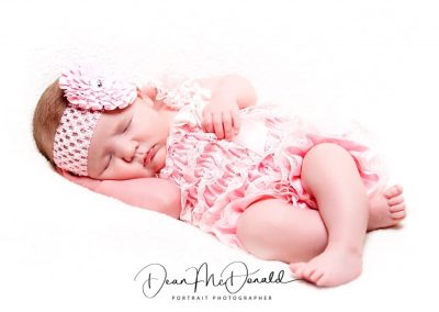 Newborn baby on a white sheet in pink dress and headband.
