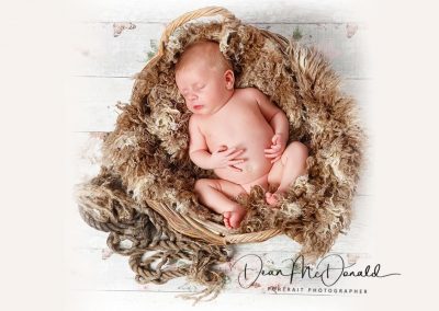 A newborn baby in a round basket covered in fur on a painted floor.
