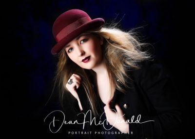 A blonde teenager with red lipstick in a black jacket with gold buttons and red bowler hat