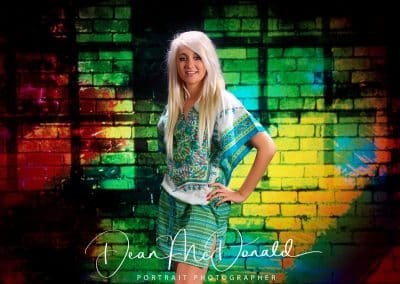A young blonde teenager in a green skirt and top posing against a multi coloured wall.