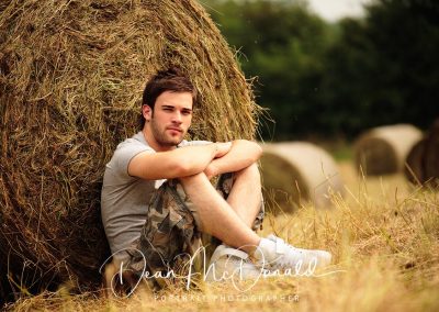 A young man can be seen sitting on hay bales in a field.